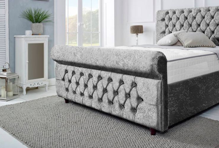 Gray tufted bed in a bedroom setting with a side table and window.